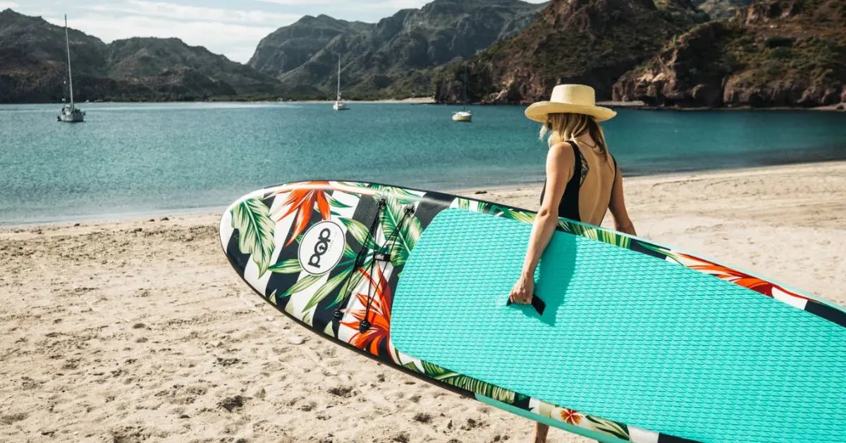 Woman carrying POP Royal Hawaiian 10'6 inflatable paddleboard with palm design on a beach.