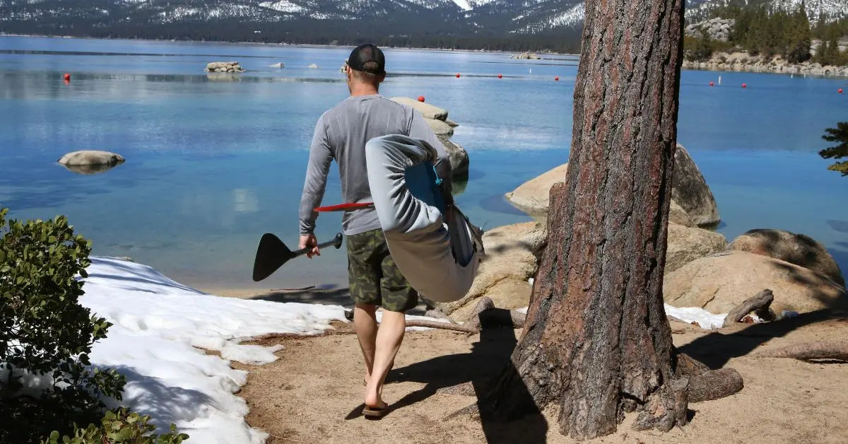 Person carrying a hard paddle board in a protective travel cover while hiking to a mountain lake