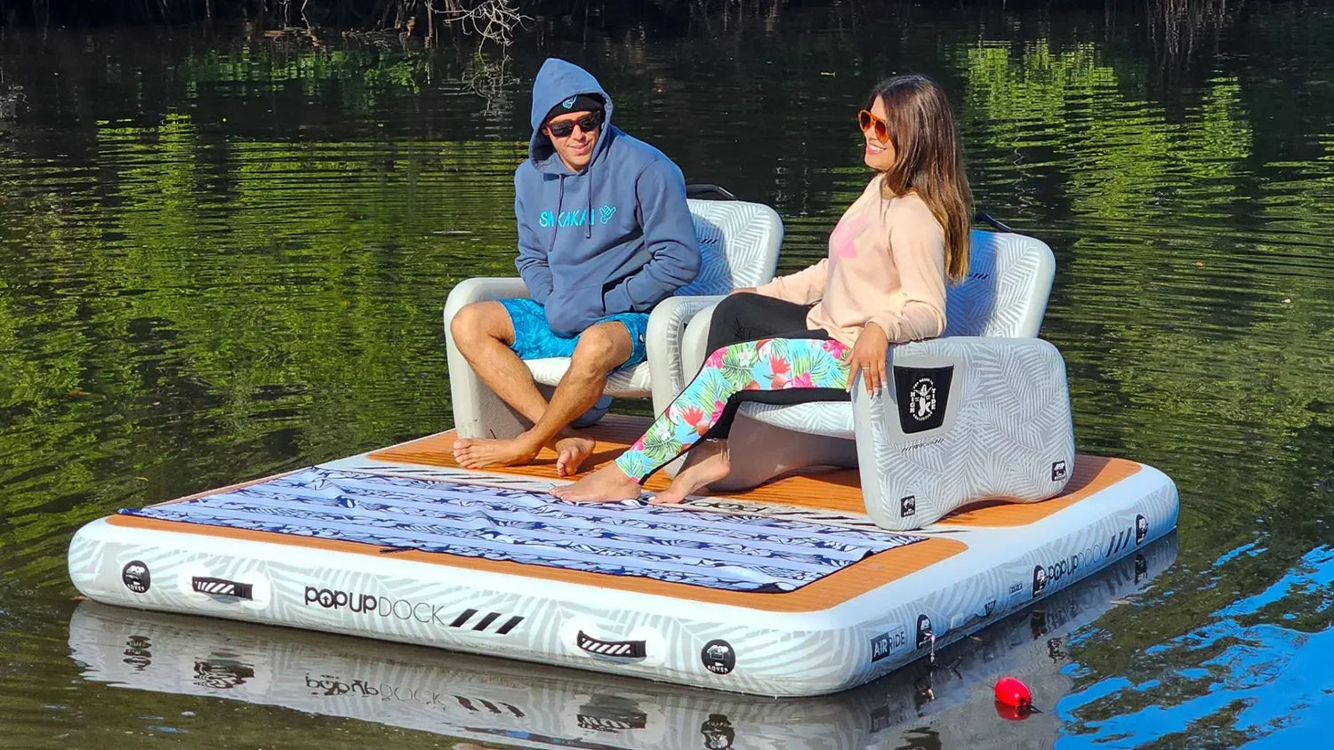 Two people relaxing on an inflatable floating dock with lounge seating on calm water.