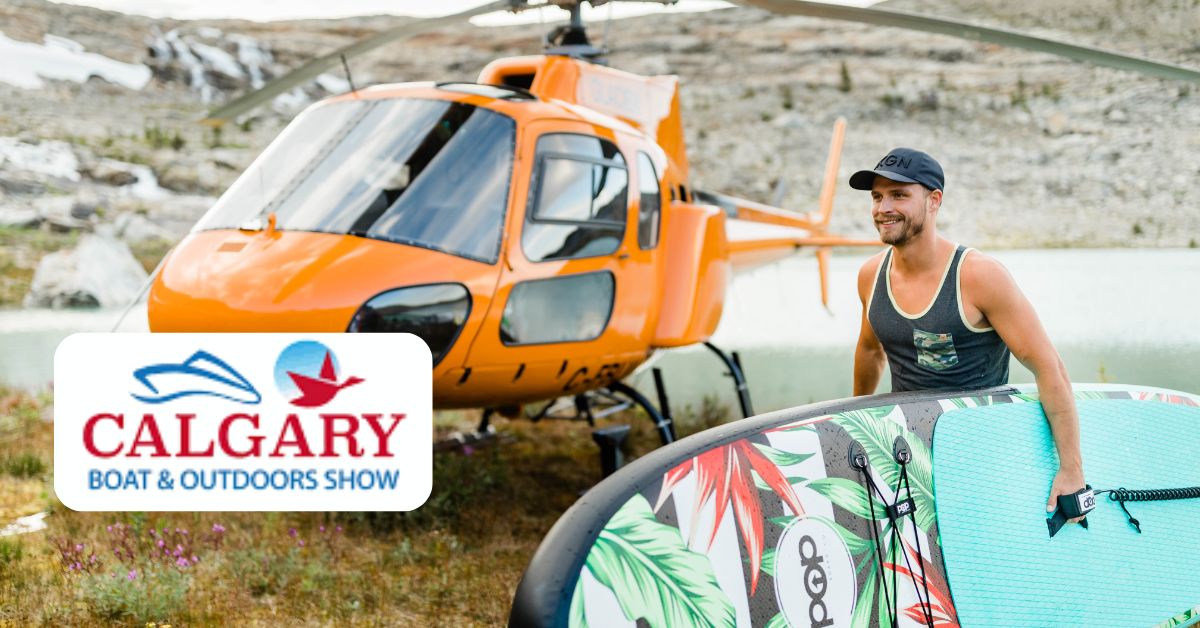 Man holding a PopBoardCo inflatable paddle board at the Calgary Boat & Outdoors Show with a helicopter in the background.