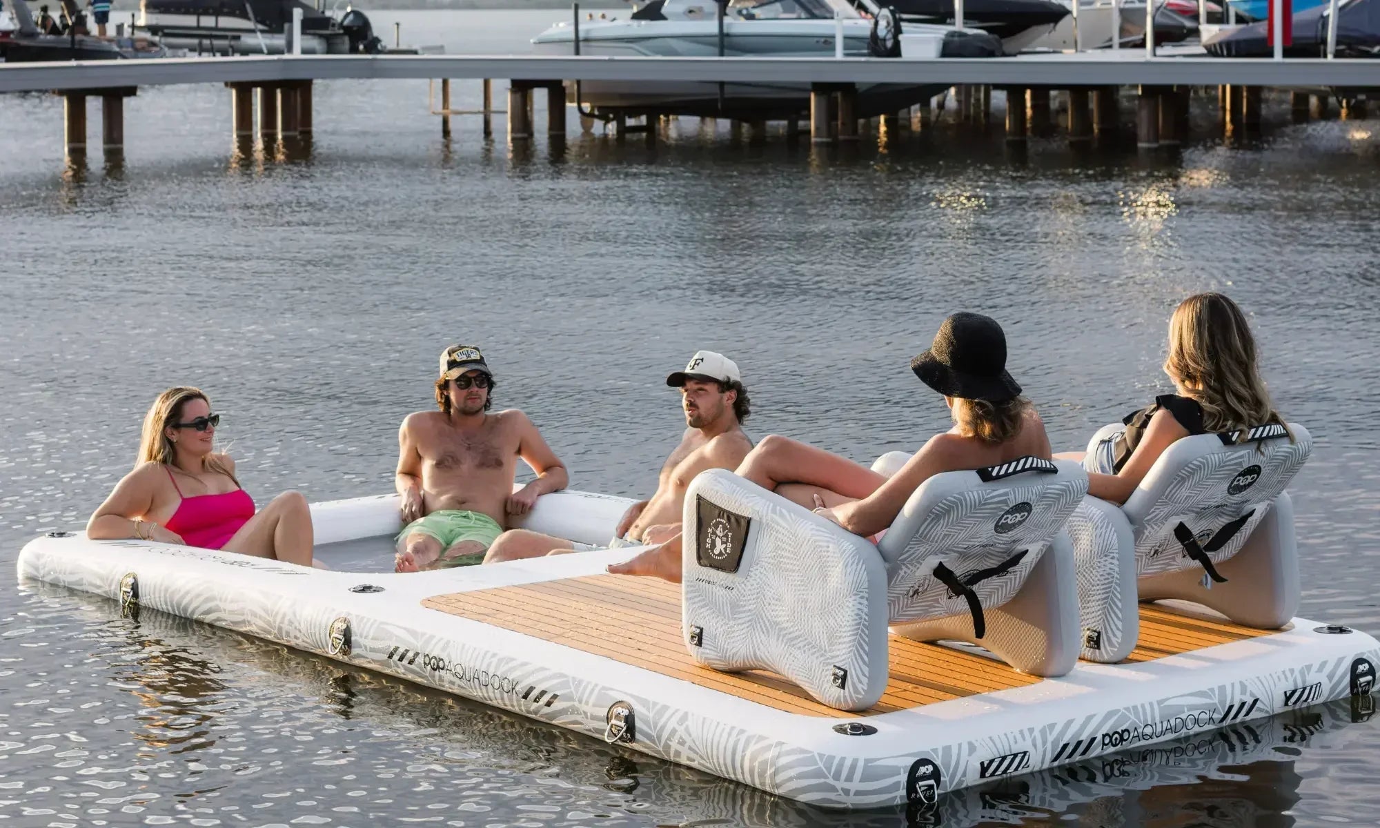 Group of people relaxing on an inflatable floating dock with lounge chairs on calm water.