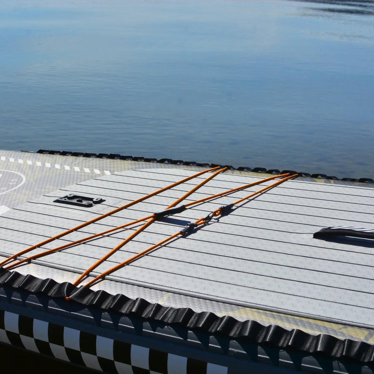 A close-up of the 11'6 El Capitan - Bomber paddleboard floating on calm water, highlighting its front deck with orange bungee cords and a checkerboard pattern along the edge.