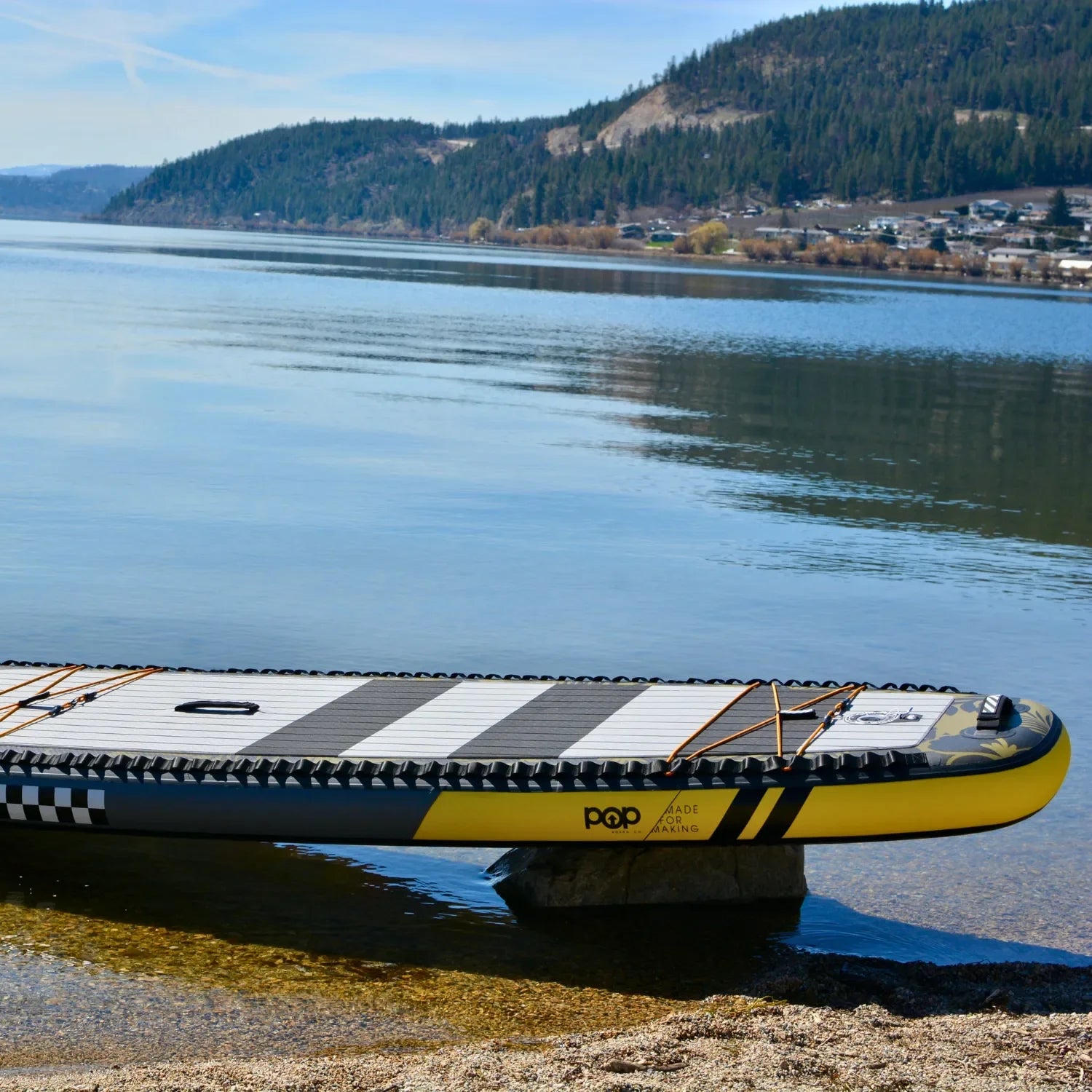 An 11'6 El Capitan - Bomber paddleboard rests on the shore of a calm lake, yellow and black against the blue sky, as forested hills and distant houses are reflected in the water.