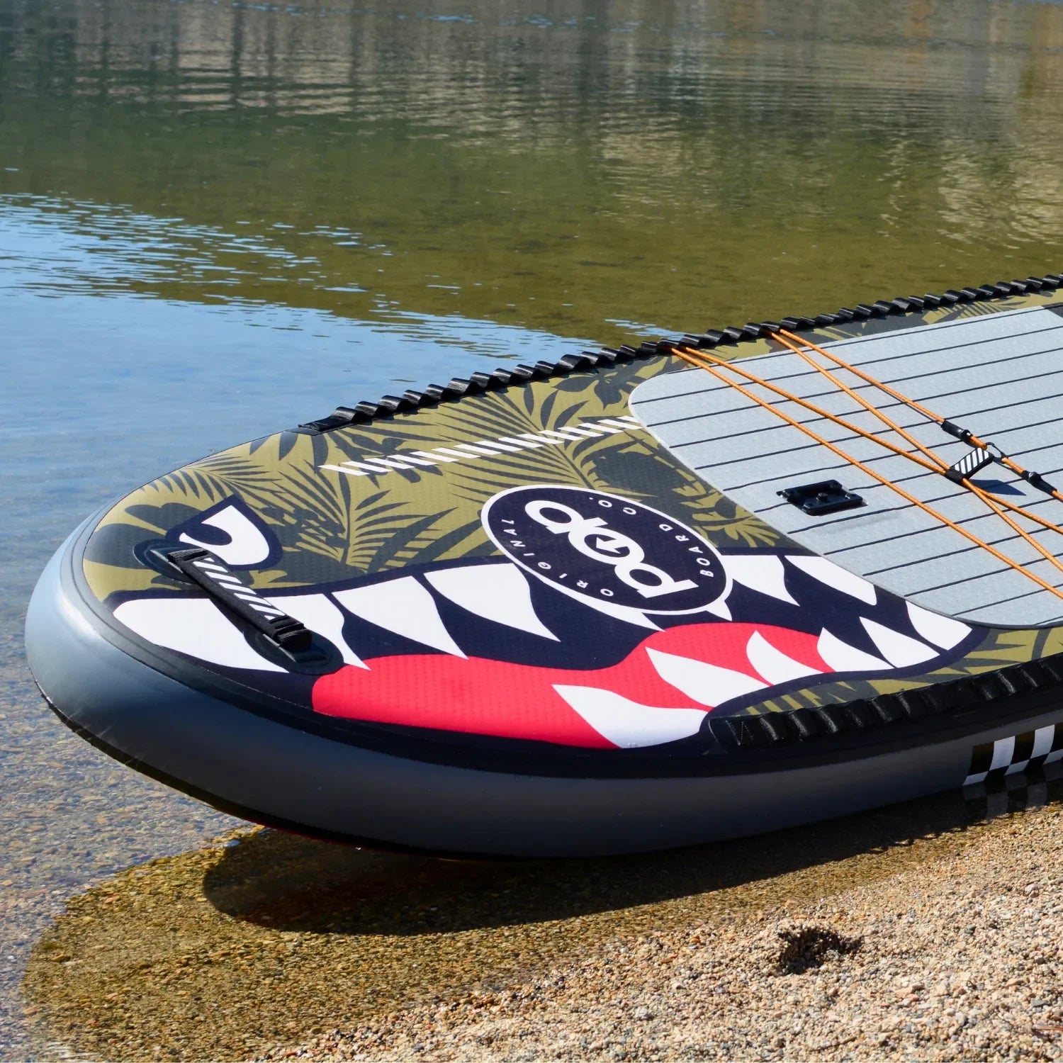 The 11'6 El Capitan - Bomber paddleboard by POP Board Co. features bold black, white, red, and khaki patterns and “pop” graphics, resting on sandy shores beside clear water. Its design offers colorful style and stability for your ride.