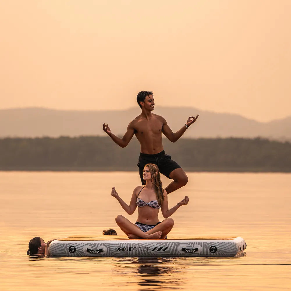 Group of friends practicing yoga on a POPUP Dock inflatable floating platform at sunset, with a man balancing on one leg and a woman sitting cross-legged in a meditative pose on the water.