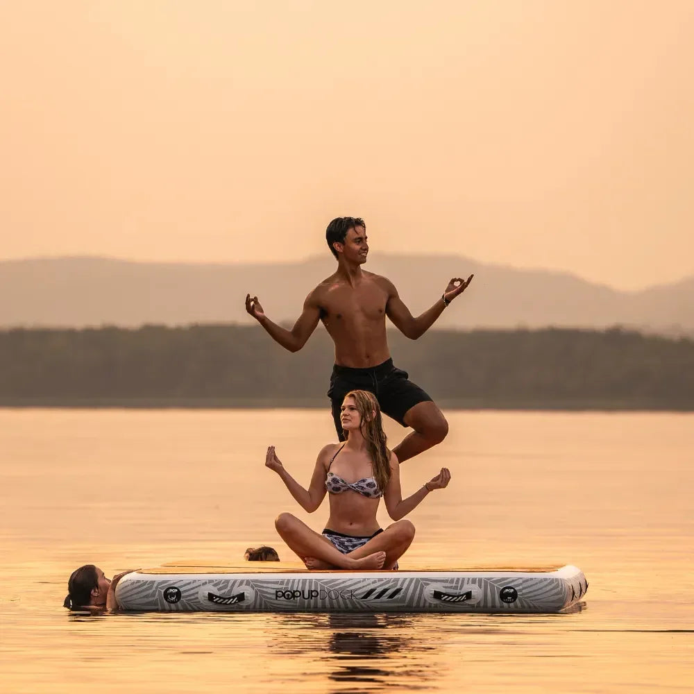 Group of friends practicing yoga on a POPUP Dock inflatable floating platform at sunset, with a man balancing on one leg and a woman sitting cross-legged in a meditative pose on the water.