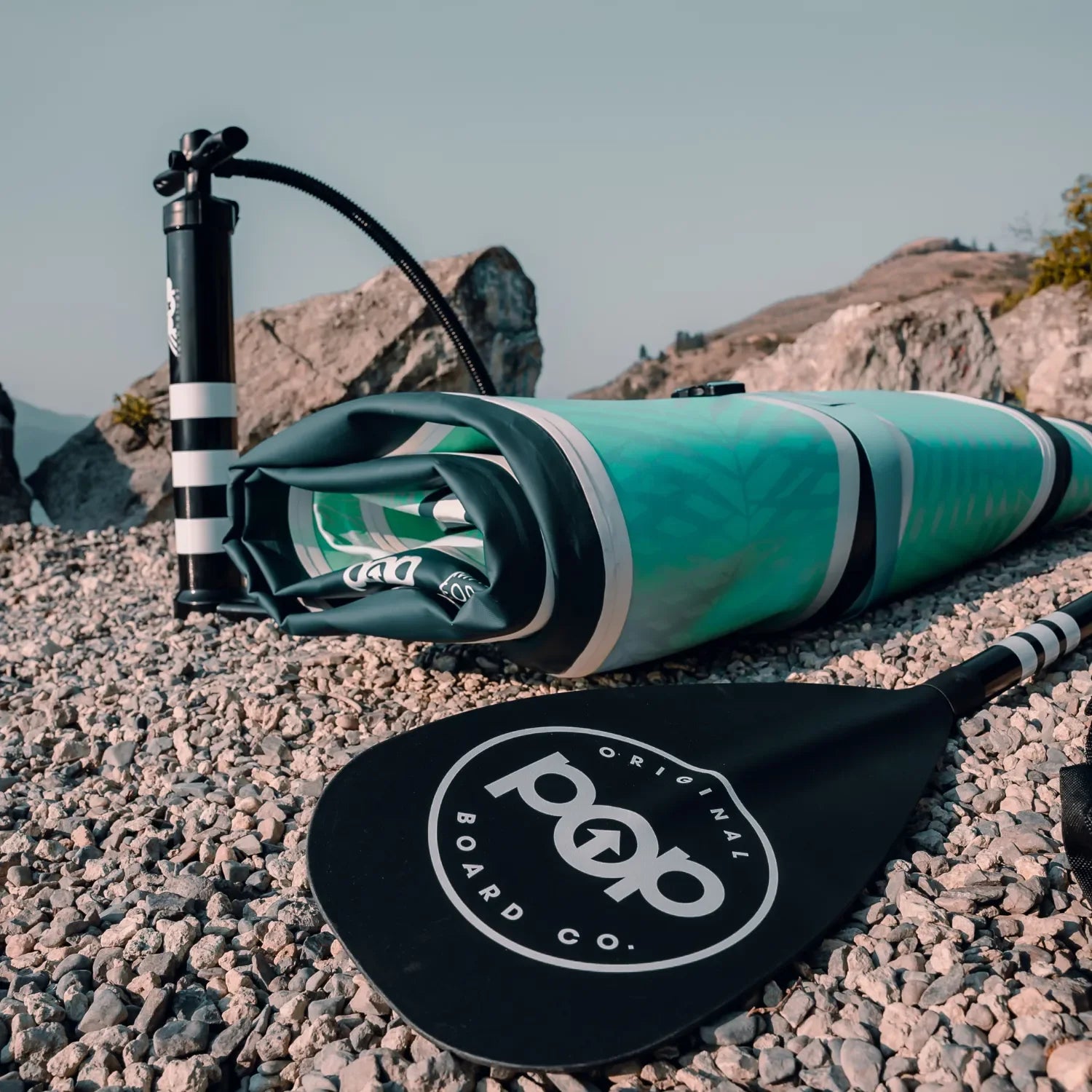A rolled-up inflatable paddleboard, a carrying bag, a POP 3 pc Aluminum Collapsible Paddle with the Original Board Co. logo, and a hand pump rest on a rocky shore with mountains in the background under a clear sky.