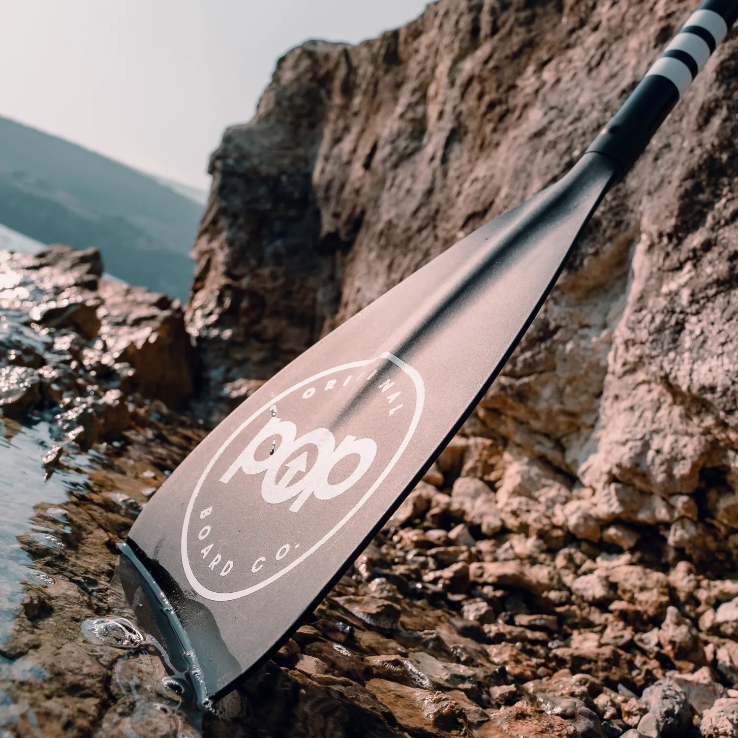 The POP 3 pc Aluminum Collapsible Paddle in black, featuring the POP BOARD CO. logo, lies on a rocky shoreline partially submerged in clear water, with sunlit rocks and blurred hills visible in the background.