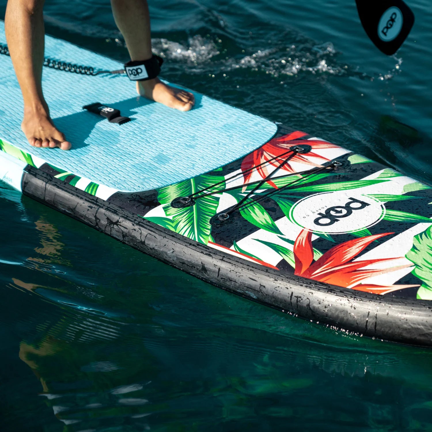 Rider standing on POP Royal Hawaiian 10'6 inflatable paddleboard with palm print design, showing stability on water.