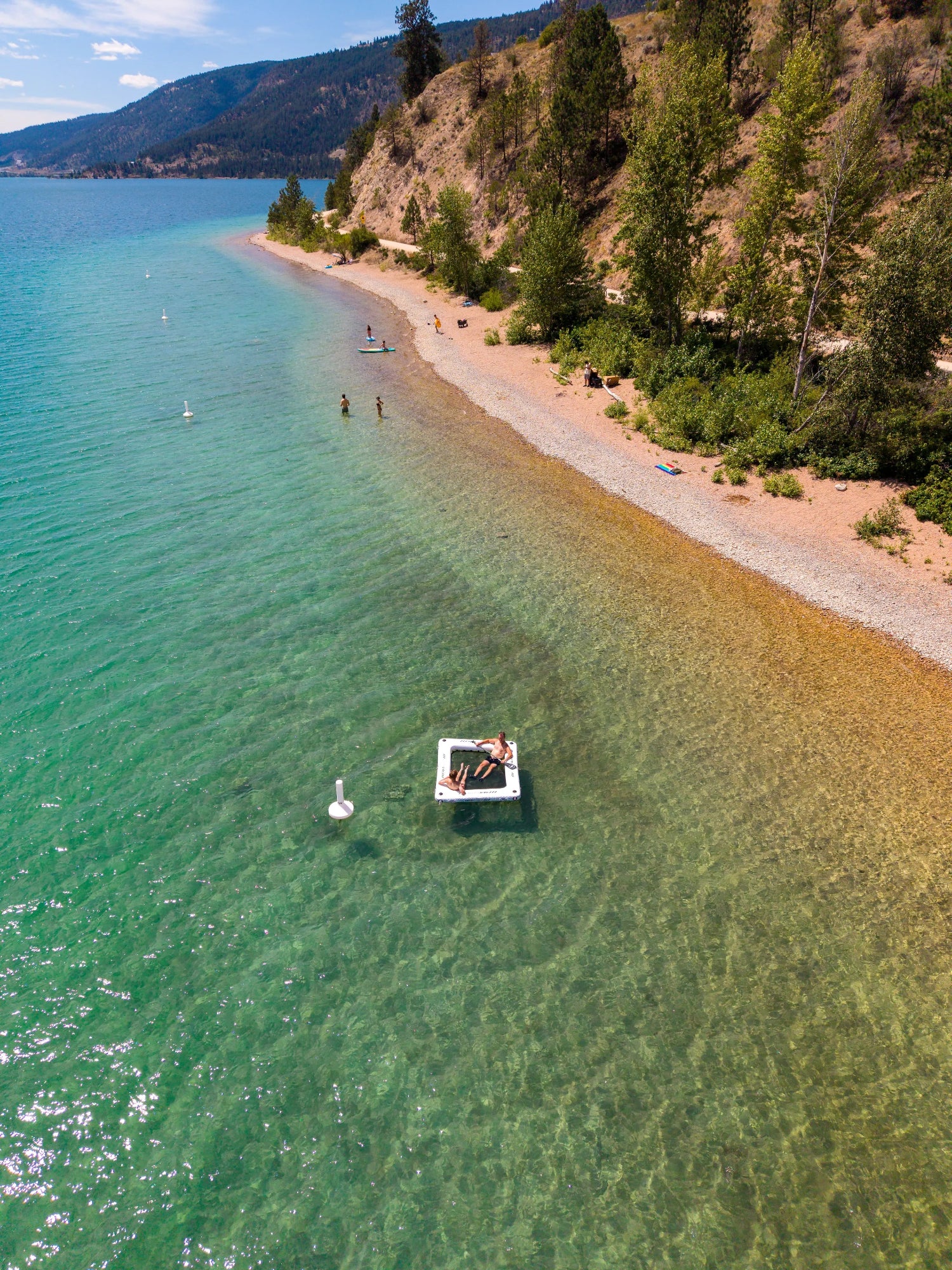 Aerial view of a clear turquoise lake beside a sandy beach and forested hillside, with a small floating platform in the shallow water near the shore.