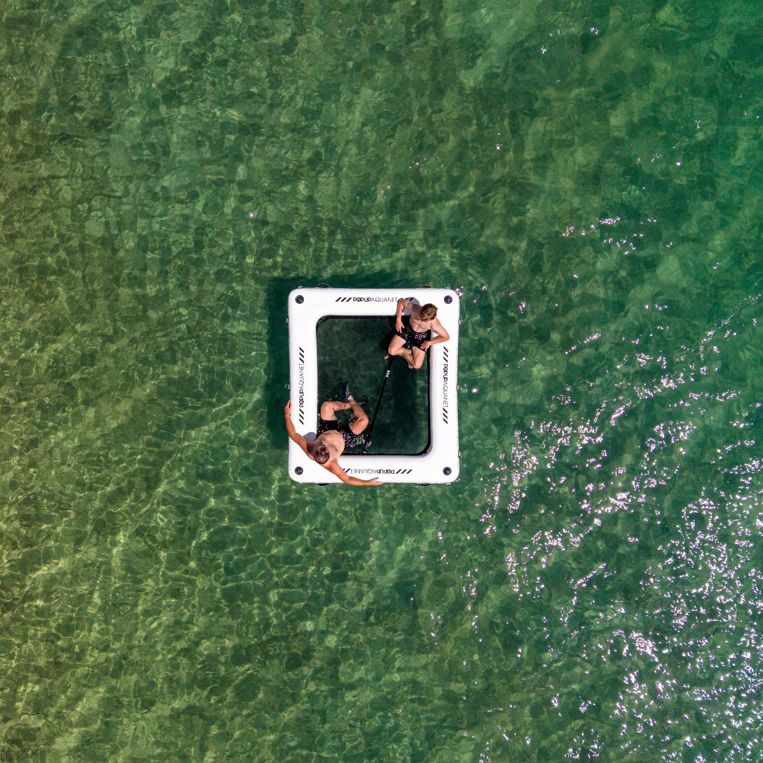An aerial view of a white square inflatable aquanet float floating on clear green lake water, with two people sitting inside and one person leaning on the side.