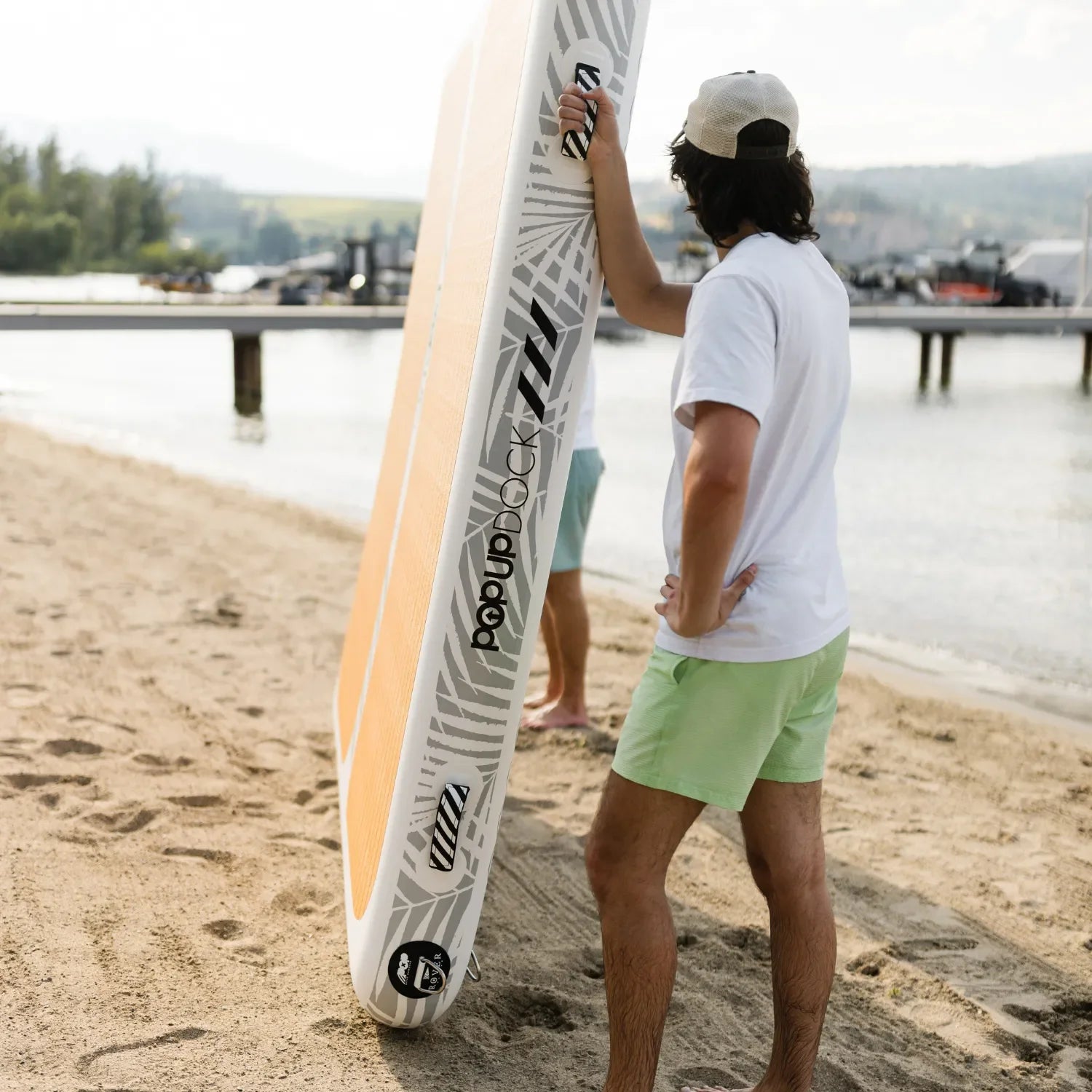 Man holding a POPUP Dock inflatable platform upright on the beach, showing its side design with traction pad and logo near the shoreline.