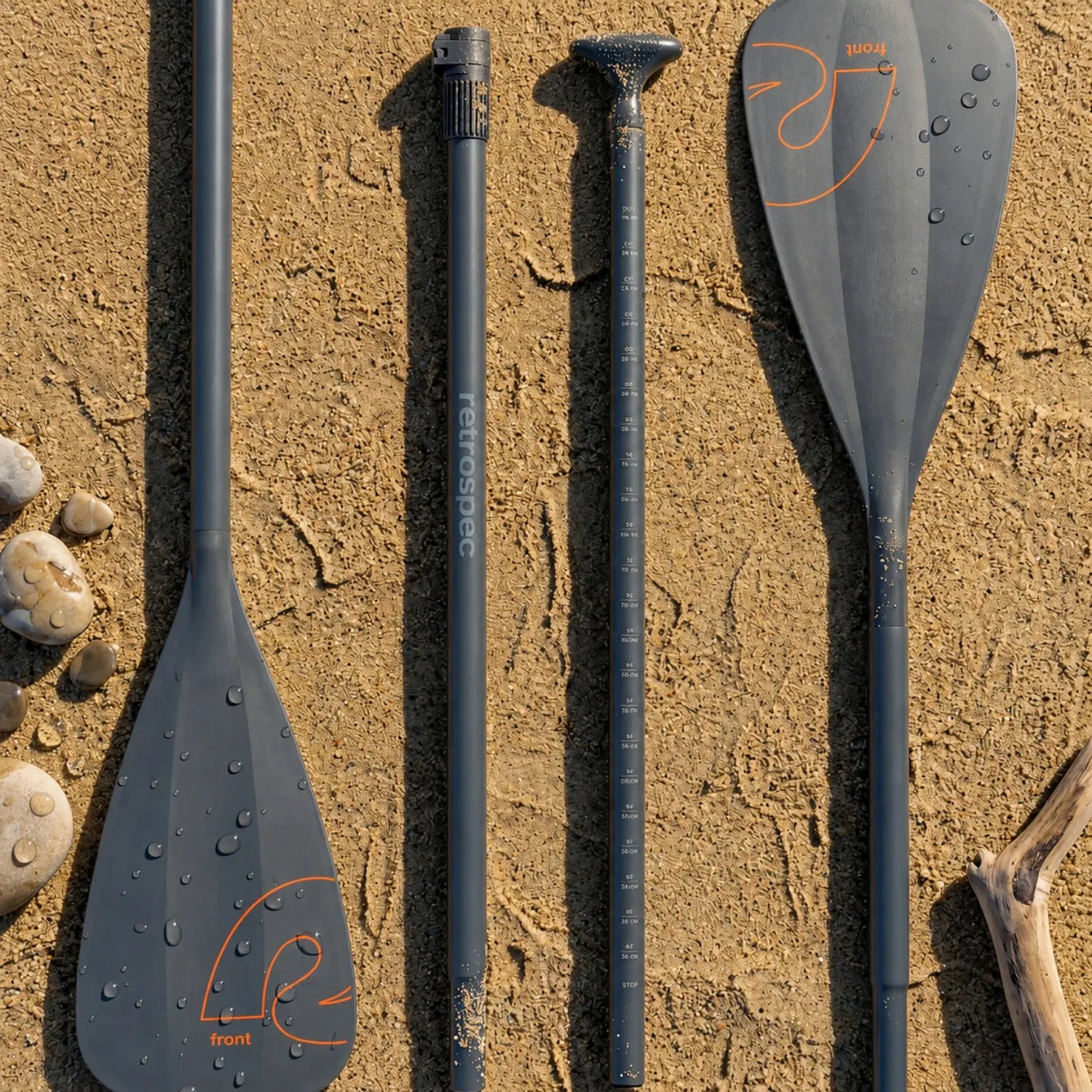 The four components of the Retrospec Weekender 2-in-1 hybrid paddle are laid out on textured beach sand, showcasing the two paddle blades and the adjustable shaft sections, complete with height markings. The dark gray pieces feature the orange 'retrospec' and 'R front' logos. Water droplets are visible on the blades, and smooth beach pebbles and a piece of driftwood frame the items, all under bright daylight.