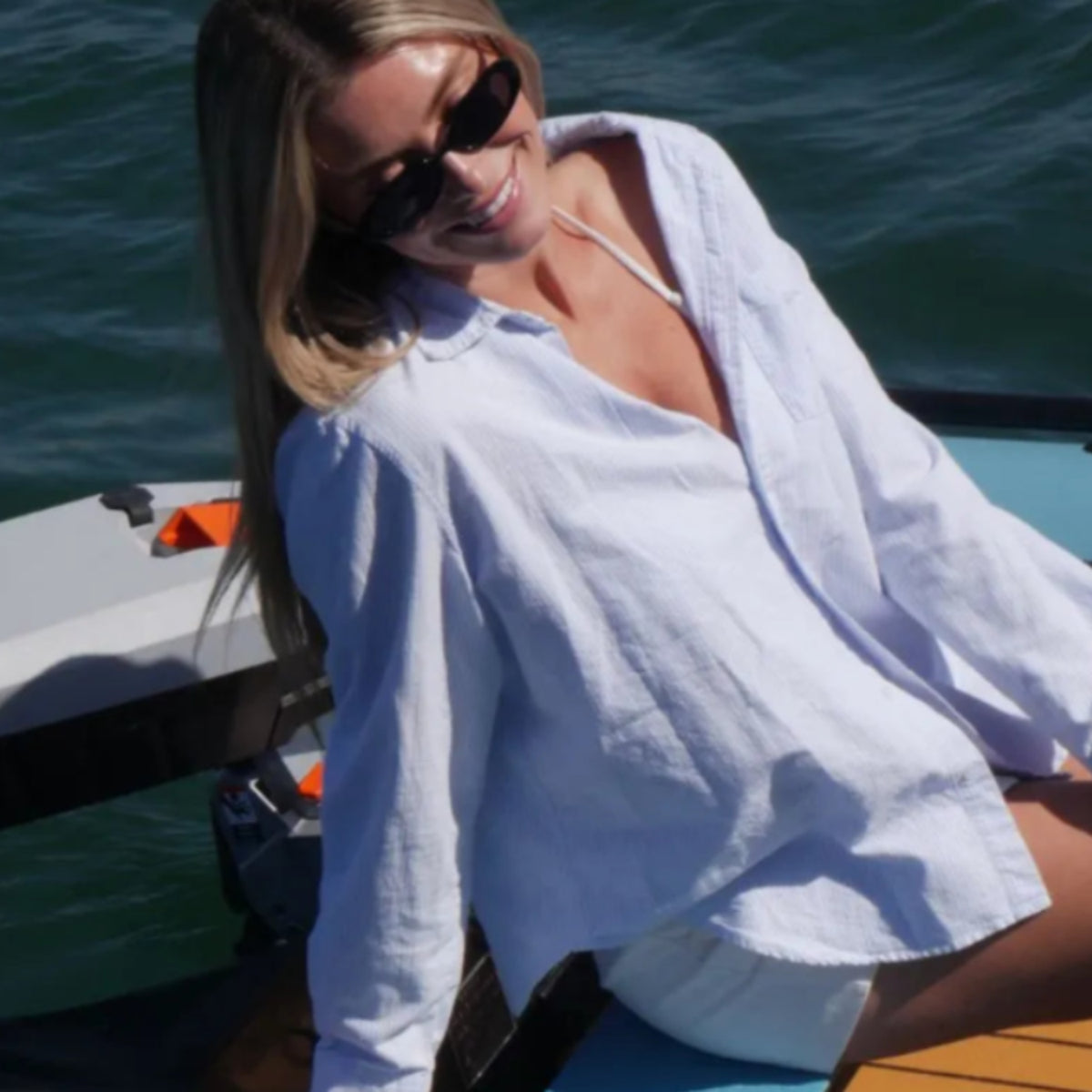 Woman sitting on a boat wearing sunglasses and a white towel, with water in the background