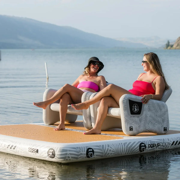 Two women in swimsuits lounging on inflatable chairs placed on a POPUP Dock floating platform, enjoying a sunny day on the water.