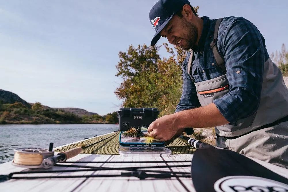 Man preparing fishing gear on an inflatable paddle board beside a calm river.