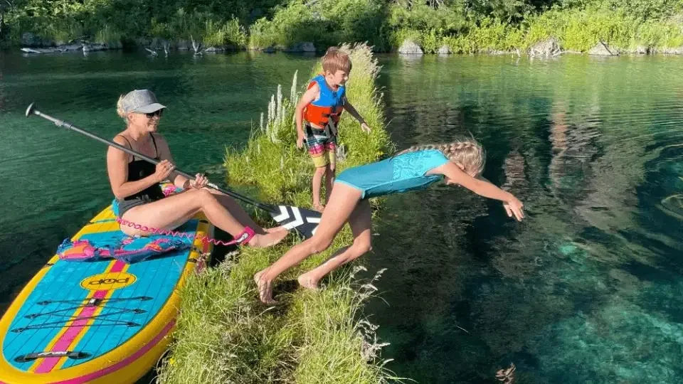 Family enjoying an inflatable paddle board on a clear lake with kids jumping into the water