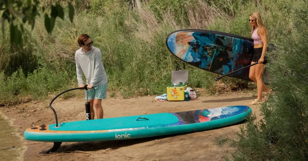 Two people inflating and carrying an inflatable paddle board on a sandy riverside