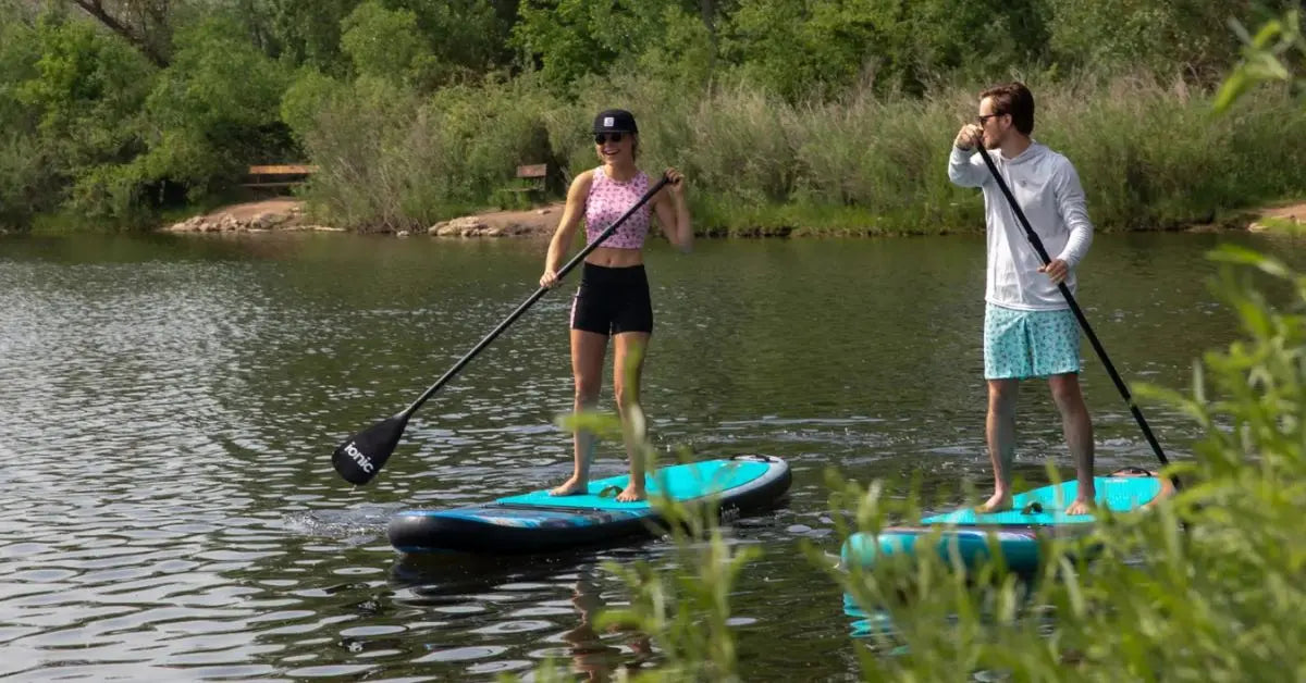 Two people paddle boarding on a calm river using inflatable paddle boards.