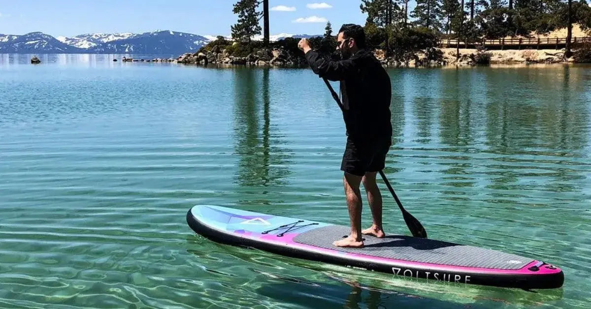 Man paddleboarding on an inflatable SUP in a clear mountain lake surrounded by pine trees and snowcapped peaks.