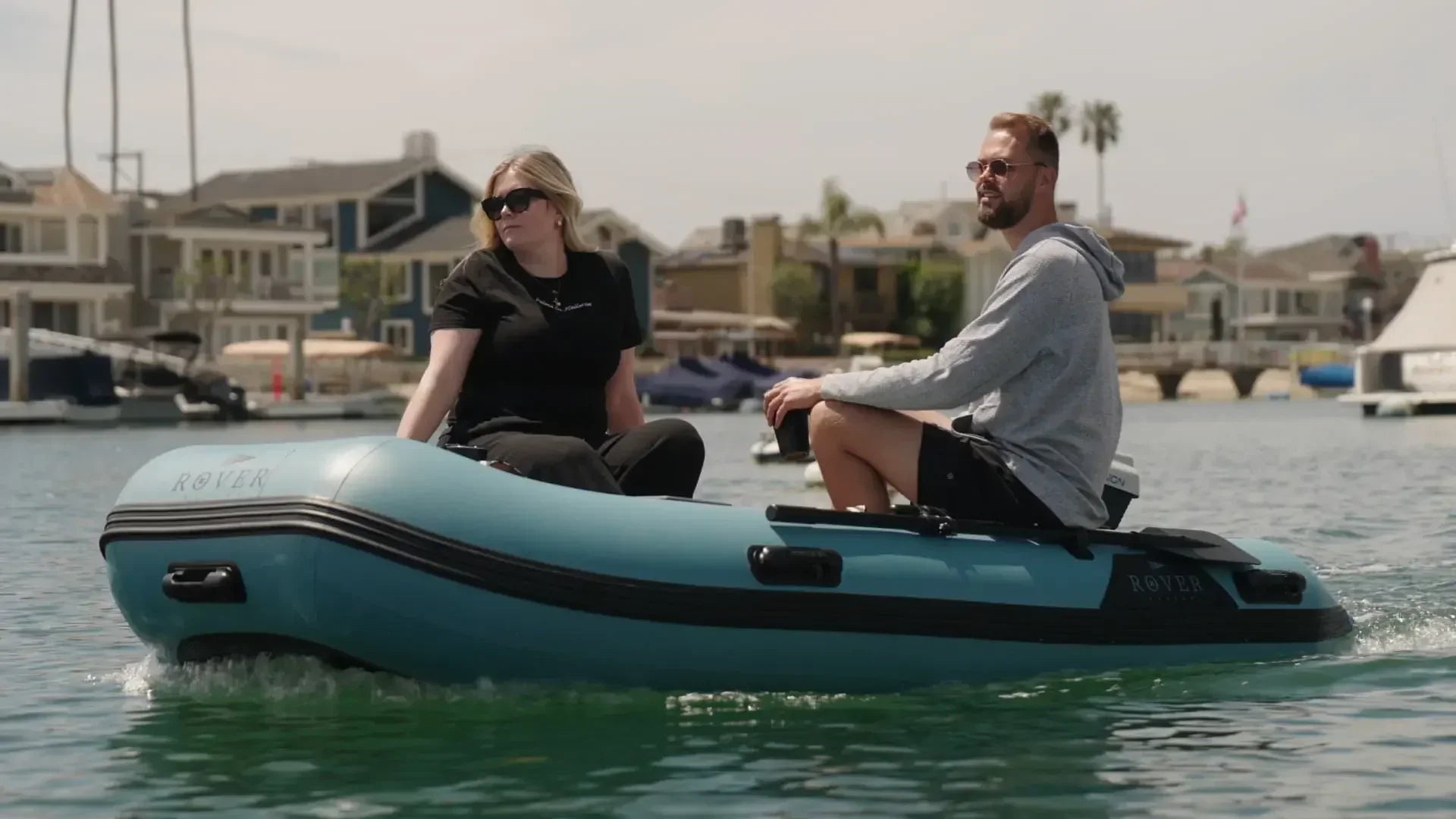 Two people riding in a Rover Marine inflatable boat on calm water near waterfront homes.