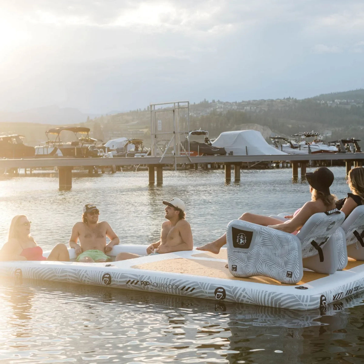 Group of friends lounging on an Aqua Dock inflatable platform with chairs, enjoying a sunny day on the water near a marina.