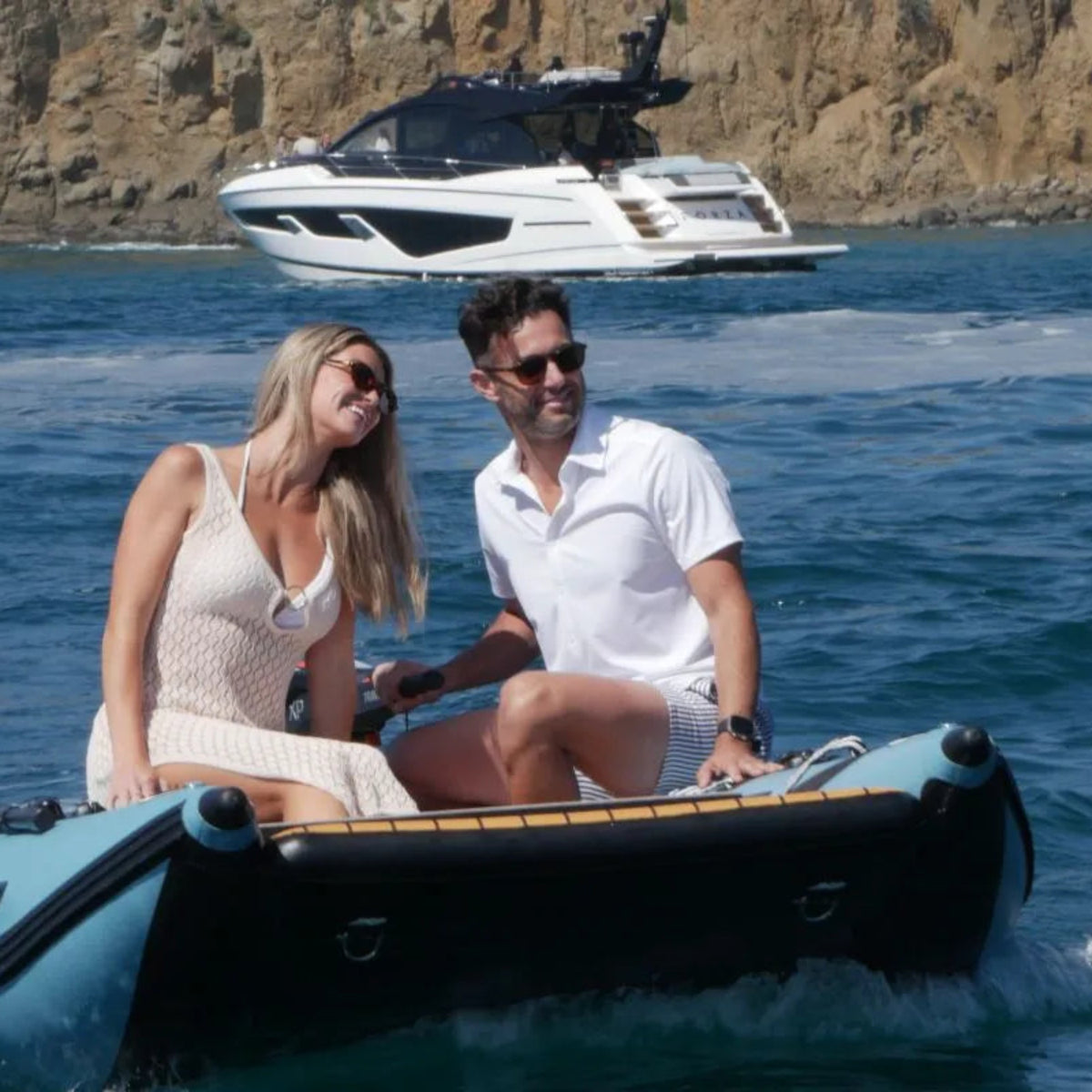 Two people in a small inflatable boat with a larger yacht in the background on a clear day.