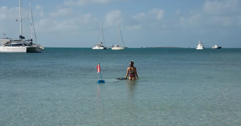 Person standing in shallow water with boats and sailboats in the background.