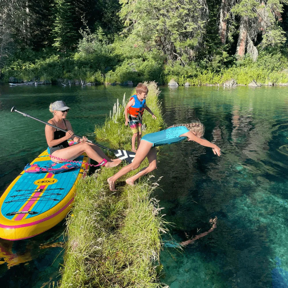 Family enjoying an inflatable paddle board on a clear lake with kids jumping into the water
