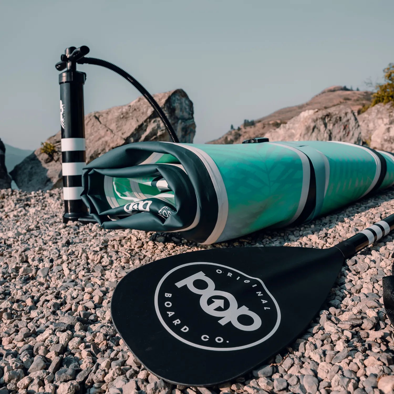 A rolled-up inflatable paddleboard, a carrying bag, a POP 3 pc Aluminum Collapsible Paddle with the Original Board Co. logo, and a hand pump rest on a rocky shore with mountains in the background under a clear sky.