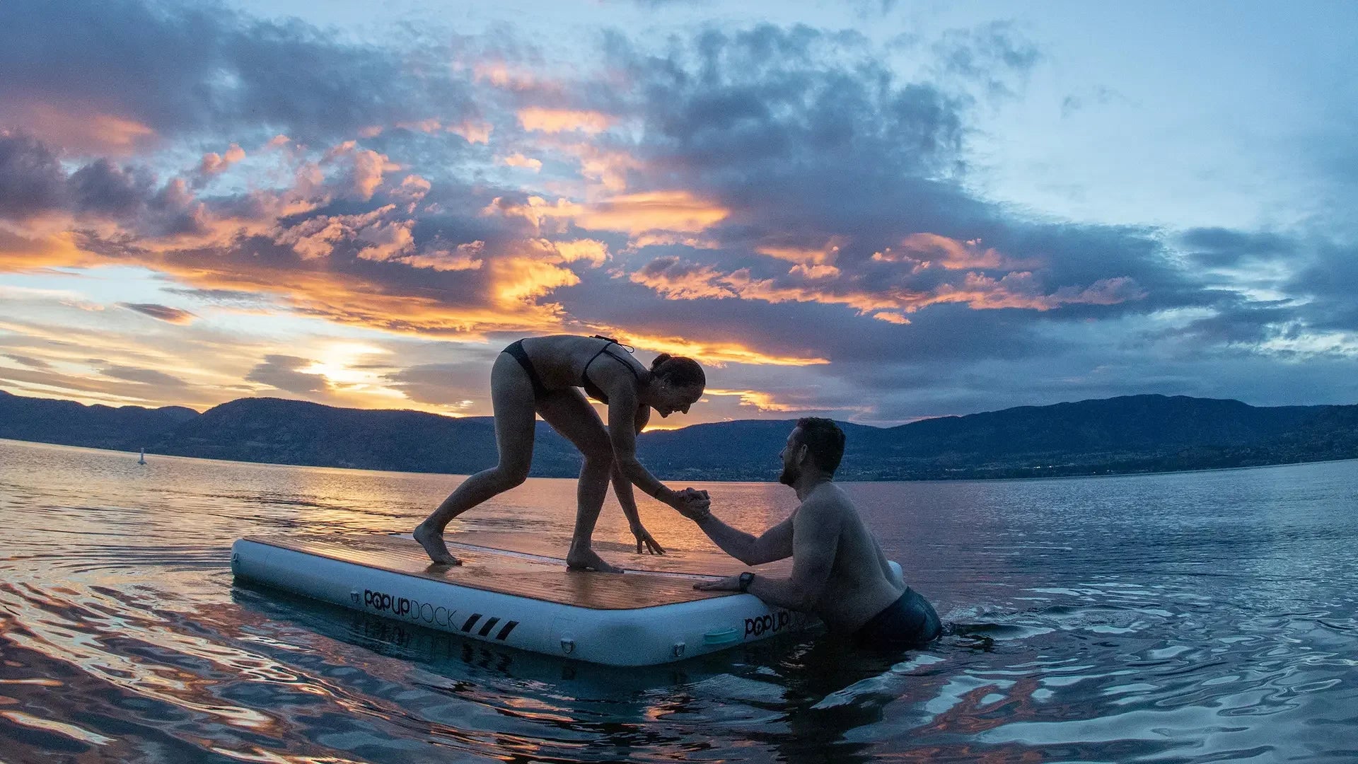 Couple enjoying an inflatable pop-up dock at sunset, with one person helping the other while floating on a calm lake