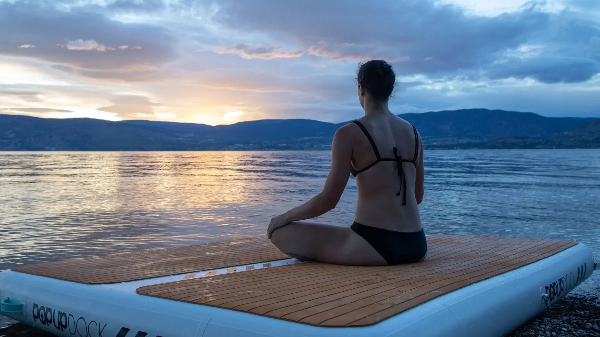 Woman meditating on an inflatable pop-up dock at sunrise with mountains and calm lake scenery in the background