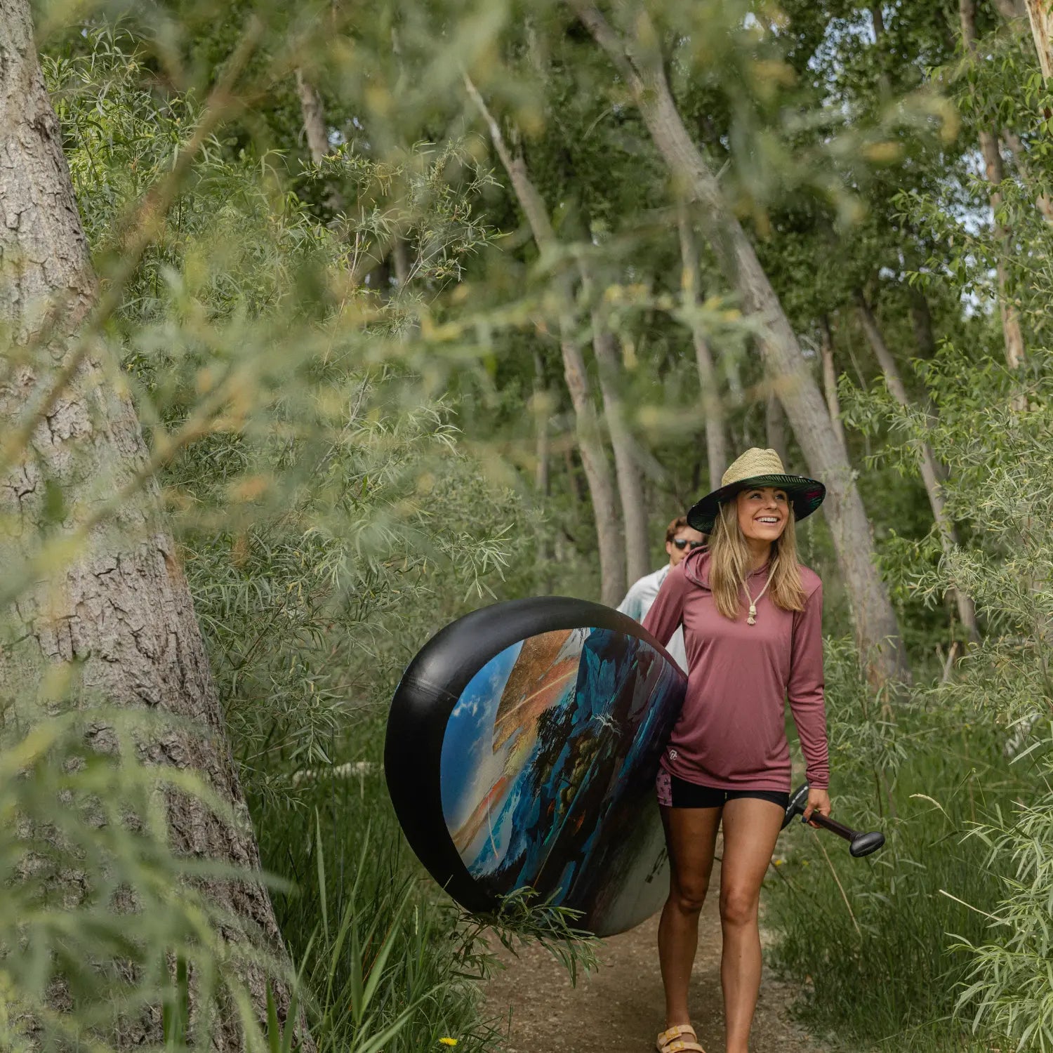 Woman carrying Ionic paddle board with Travis Parr limited edition graphics through forest trail.