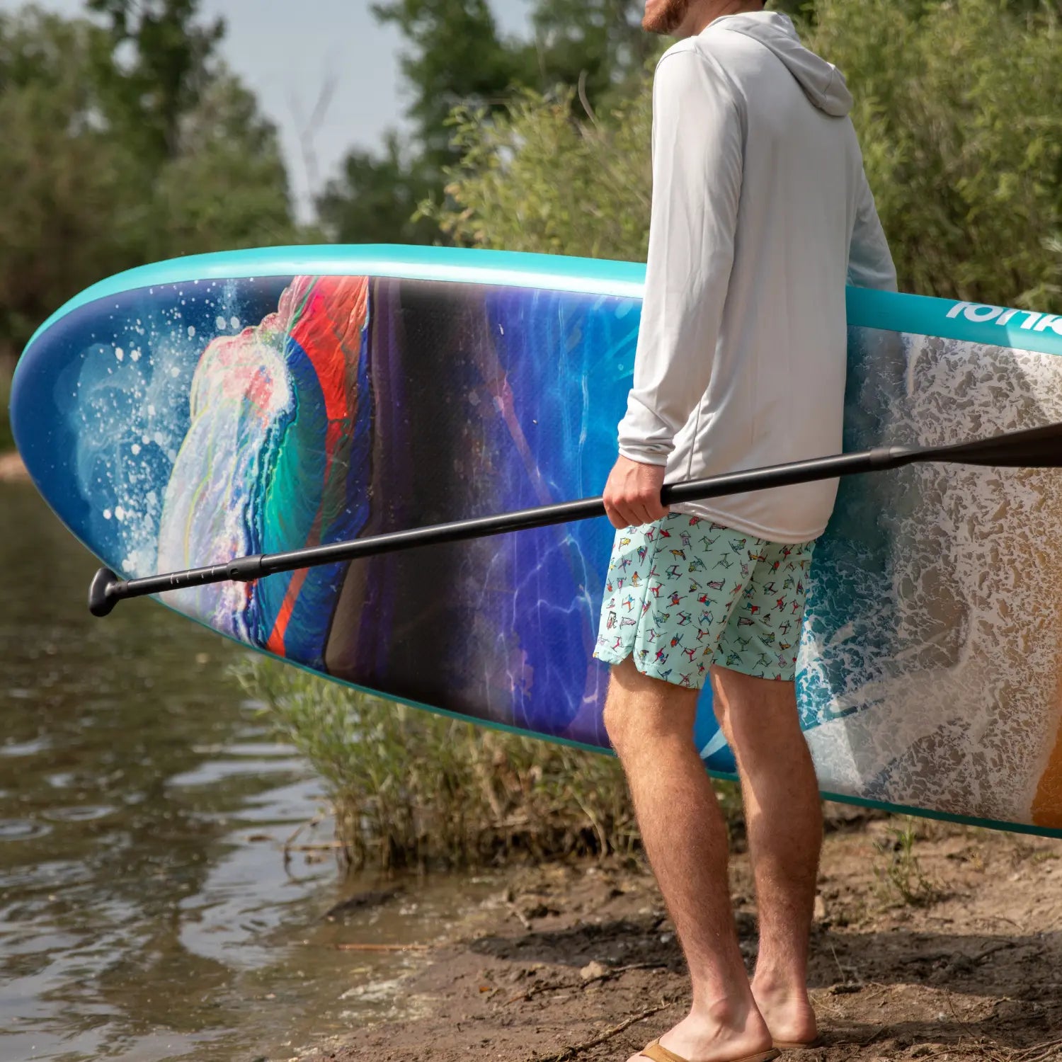 Close-up of man holding Ionic paddle board with Travis Parr colorful artwork design by the water.