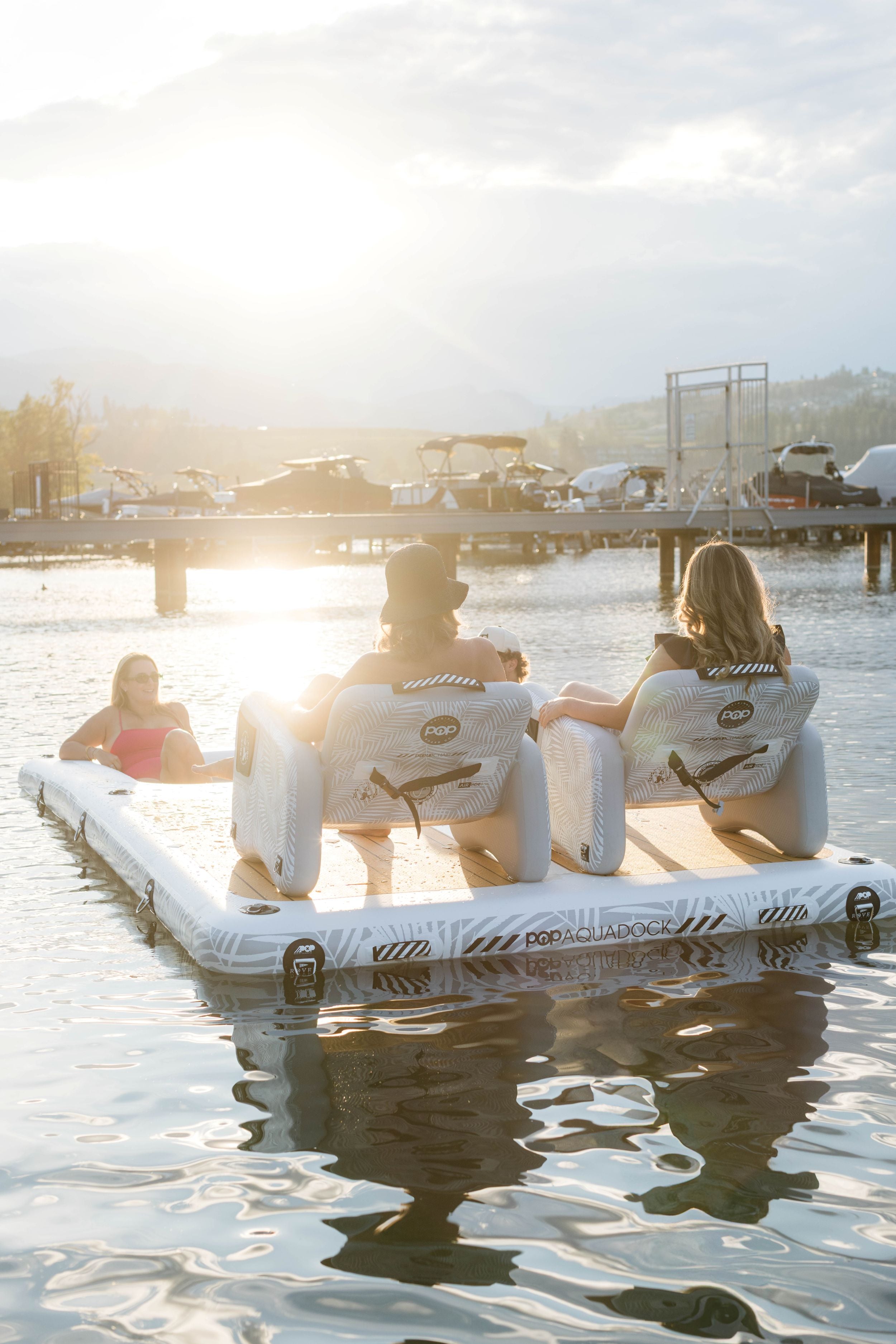 Group relaxing on POP Aqua Dock with inflatable chairs, enjoying the sunset on the water near a marina.