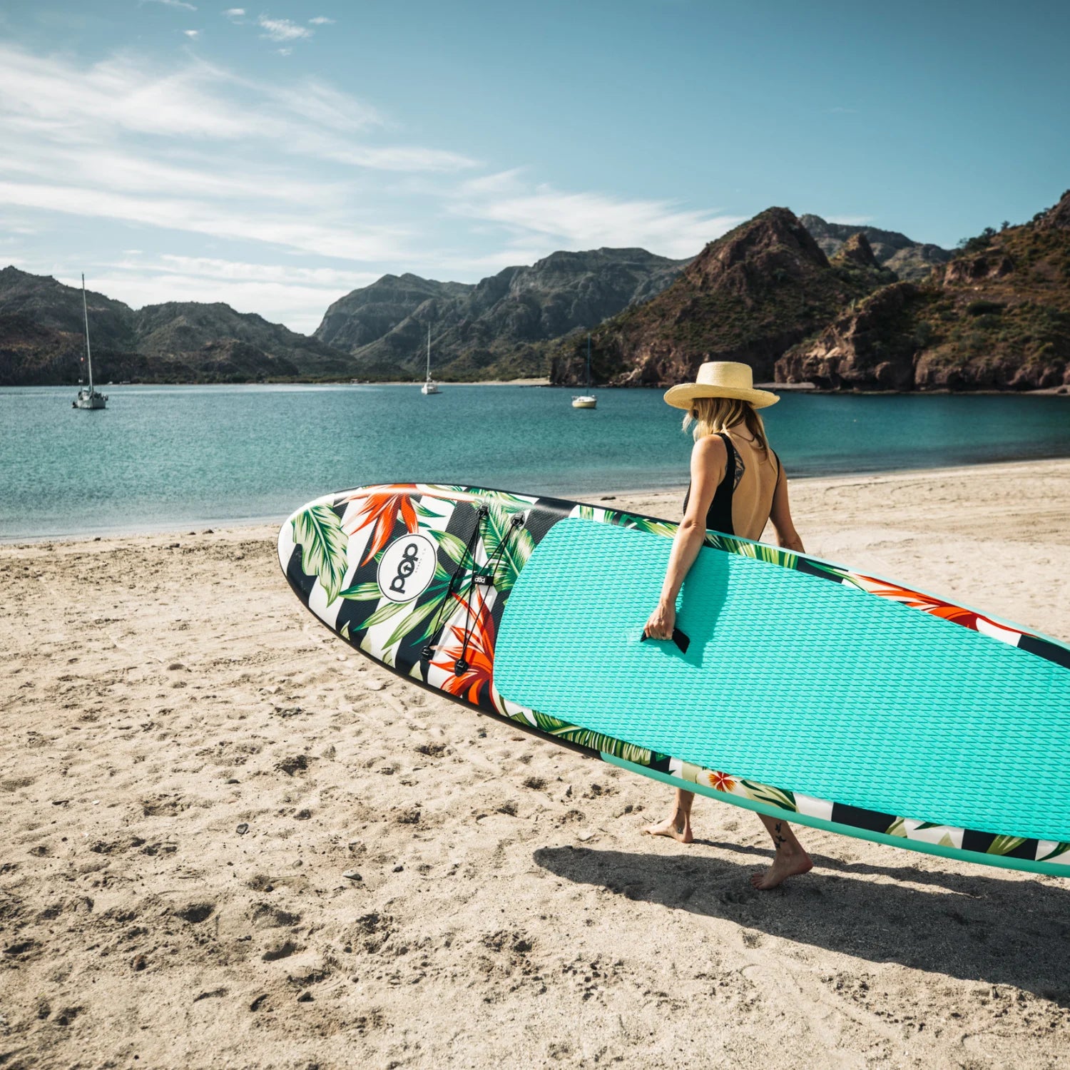 Woman carrying POP Royal Hawaiian 10'6 inflatable paddleboard with palm design on a beach.