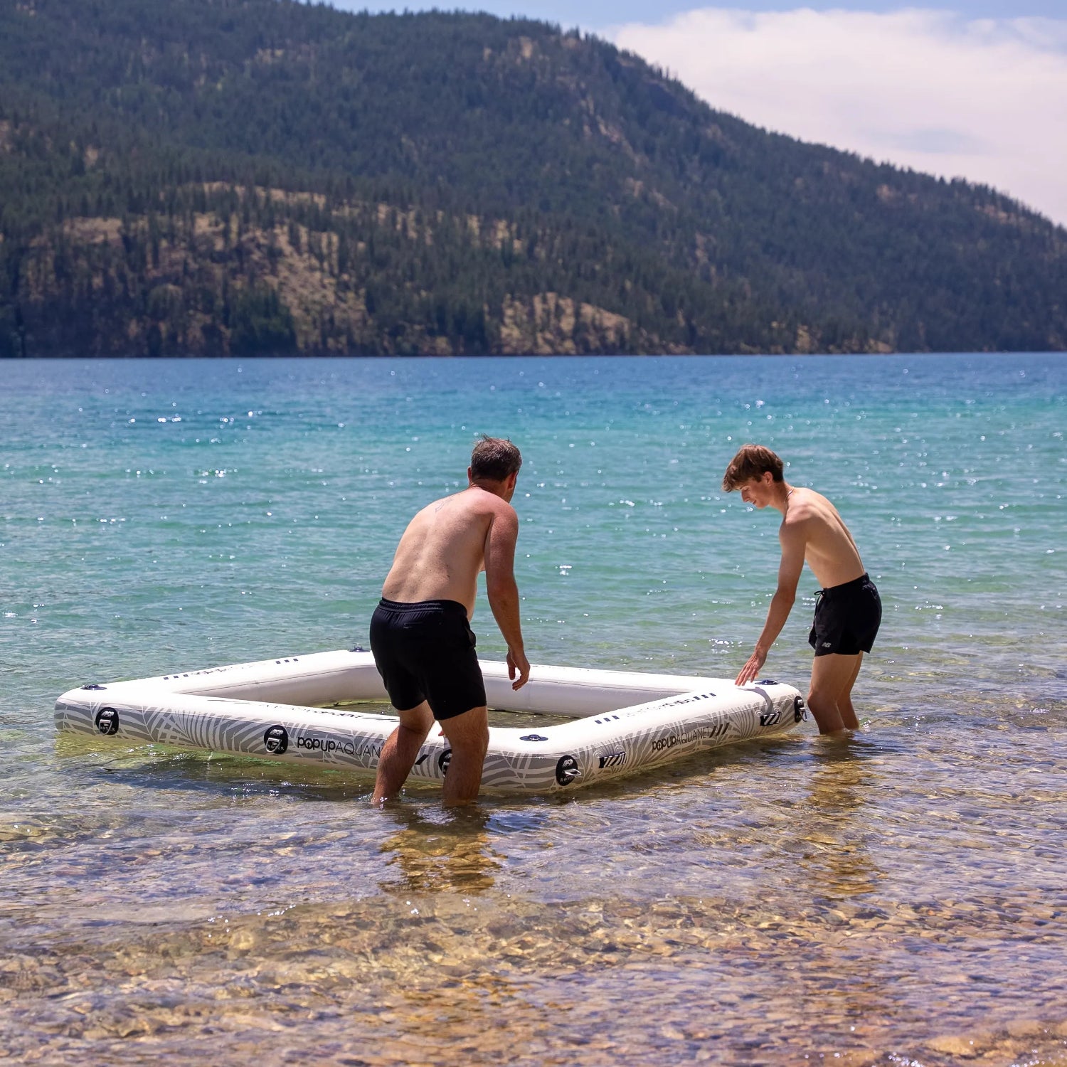 Two men standing in shallow turquoise water near a rocky shoreline, assembling a large white rectangular inflatable aquanet with black markings. A forested mountain and blue sky are visible in the background.