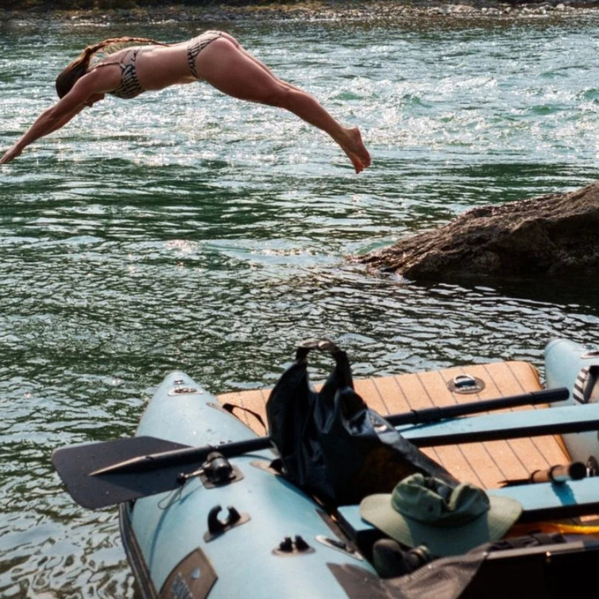 A woman jumps into a river from above, with a light blue inflatable raft and paddles in the foreground on the water.