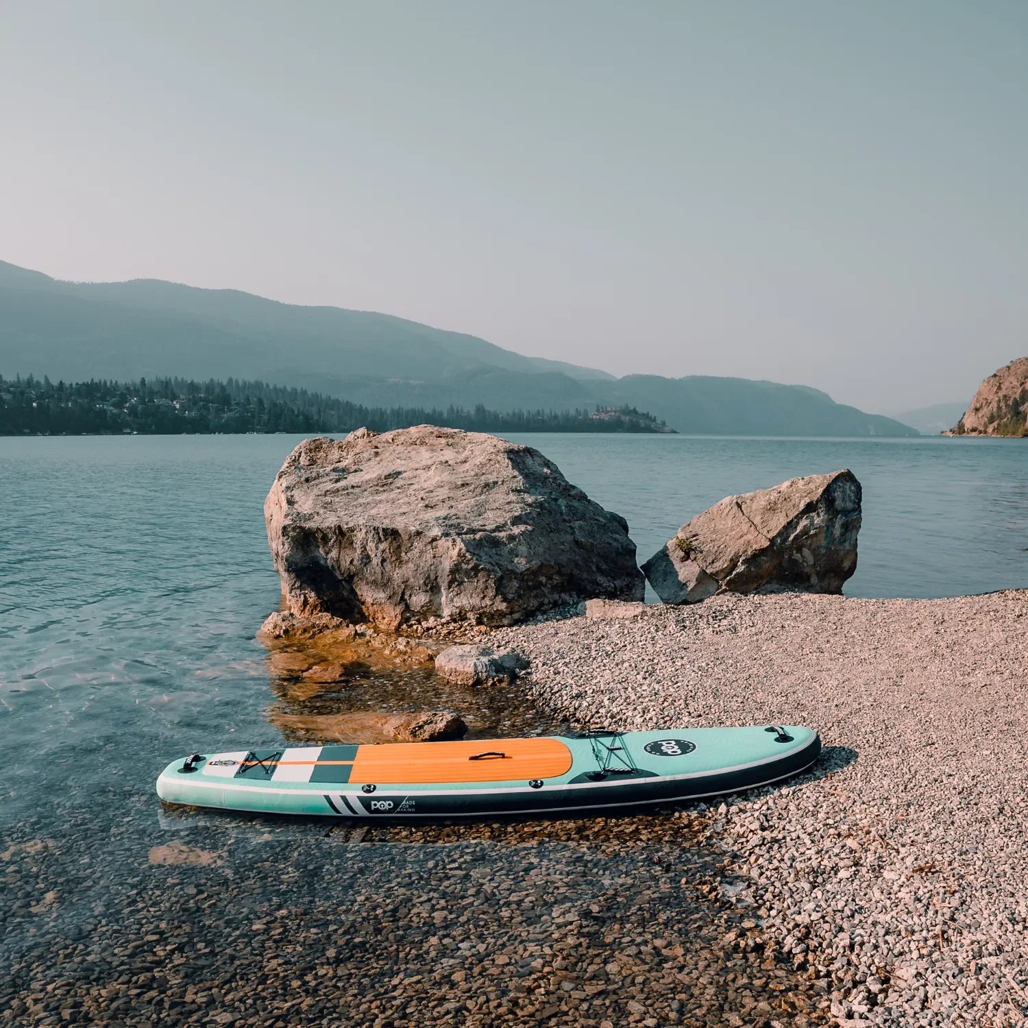 POP Yacht Hopper 11'0 inflatable paddleboard in teak blue and mint resting by rocky shoreline.